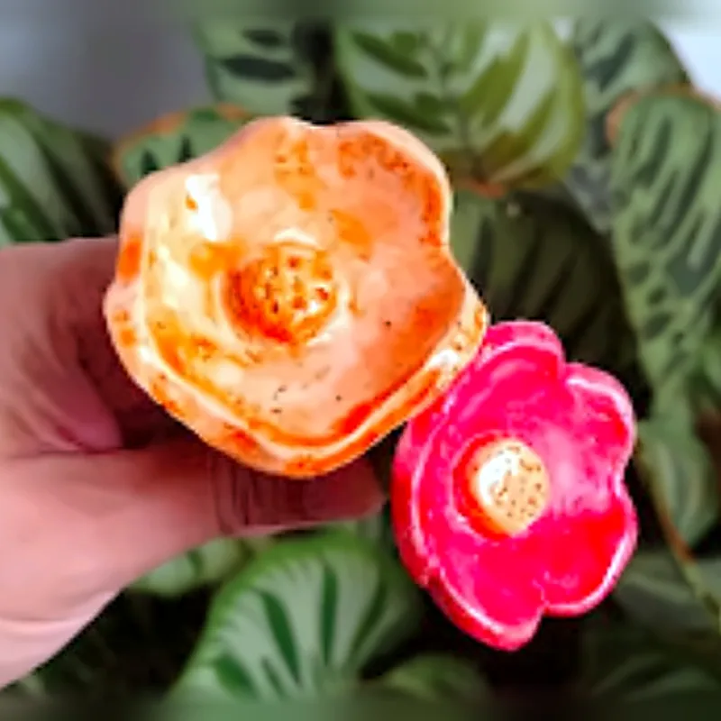 Dos flores de cerámica, una naranja y una rosa, sostenidas frente a una planta